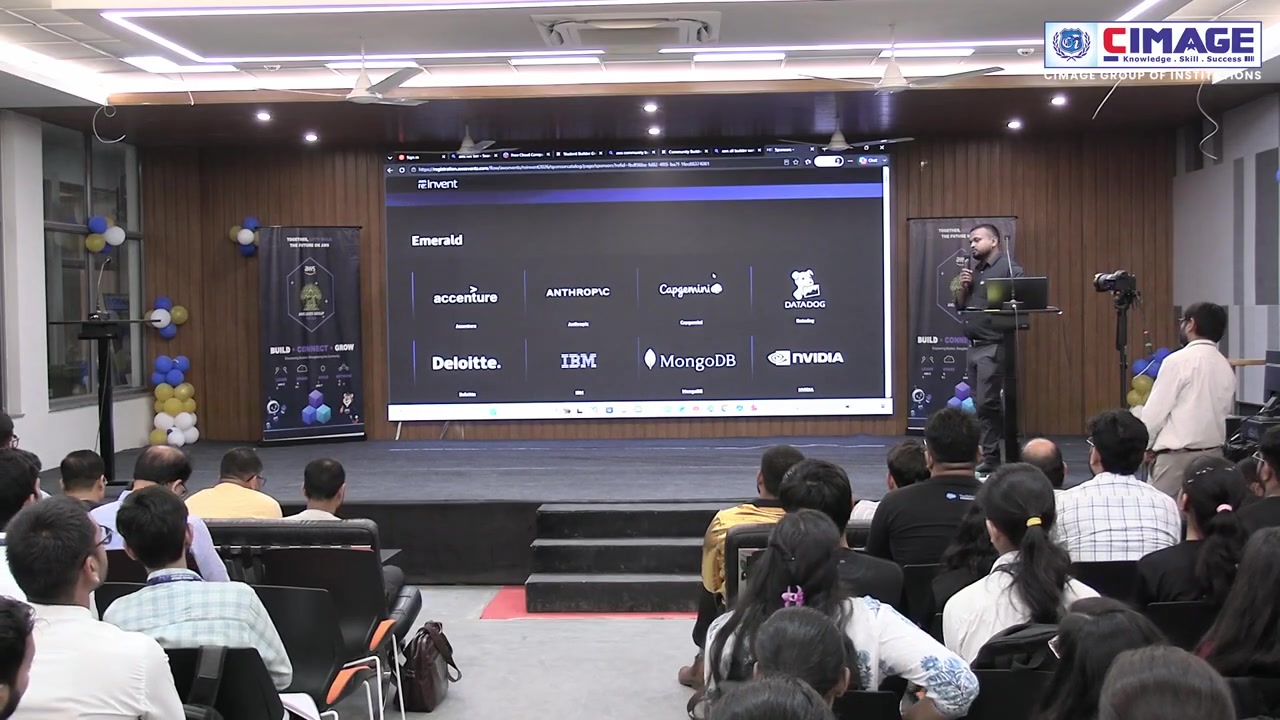 Audience of students seated in a hall facing a stage with screen.