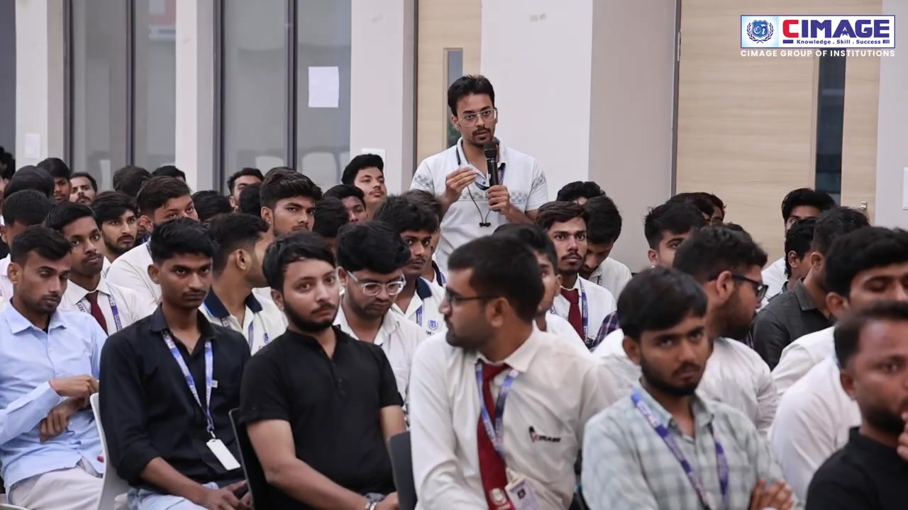 A large group of students are seated attentively in an auditorium, with one student standing and speaking.
