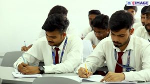 Several students are seated at desks in a classroom, attentively writing in notebooks.