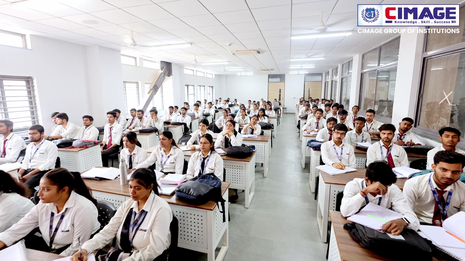 Wide-angle view of a large, brightly lit classroom filled with numerous students seated at desks.