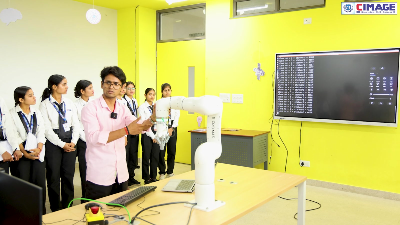 A group of students watch a robotic arm demonstration in a brightly lit classroom.