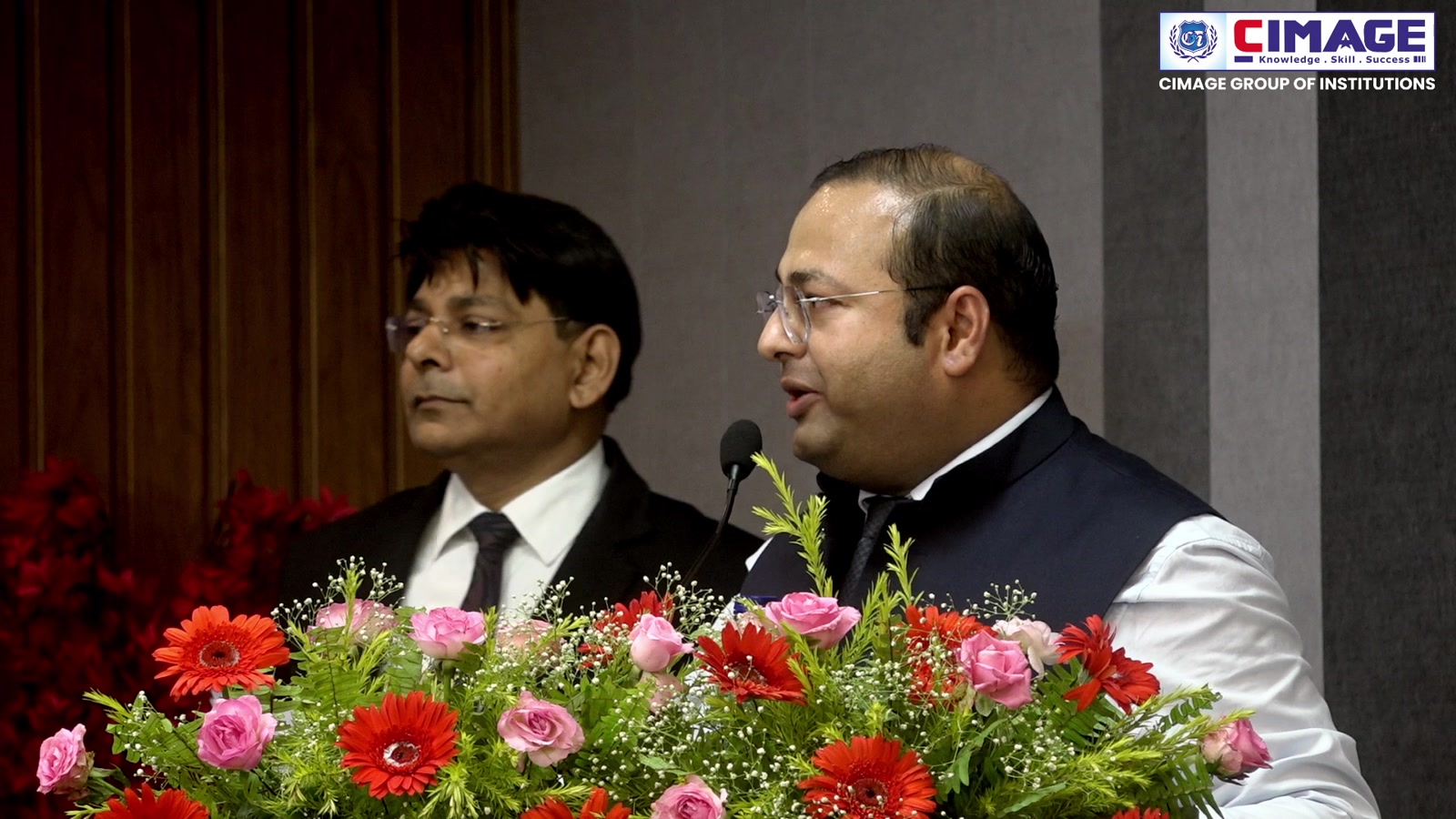 Two speakers stand behind a podium decorated with flowers at an institutional event.